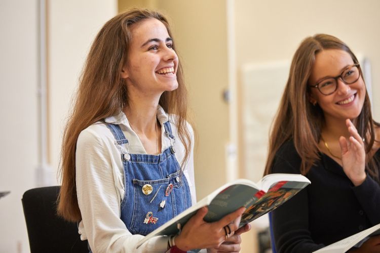 A girl with brown hair and wearing blue dungarees and a white shirt holds out a book. She has a big smile across her face.